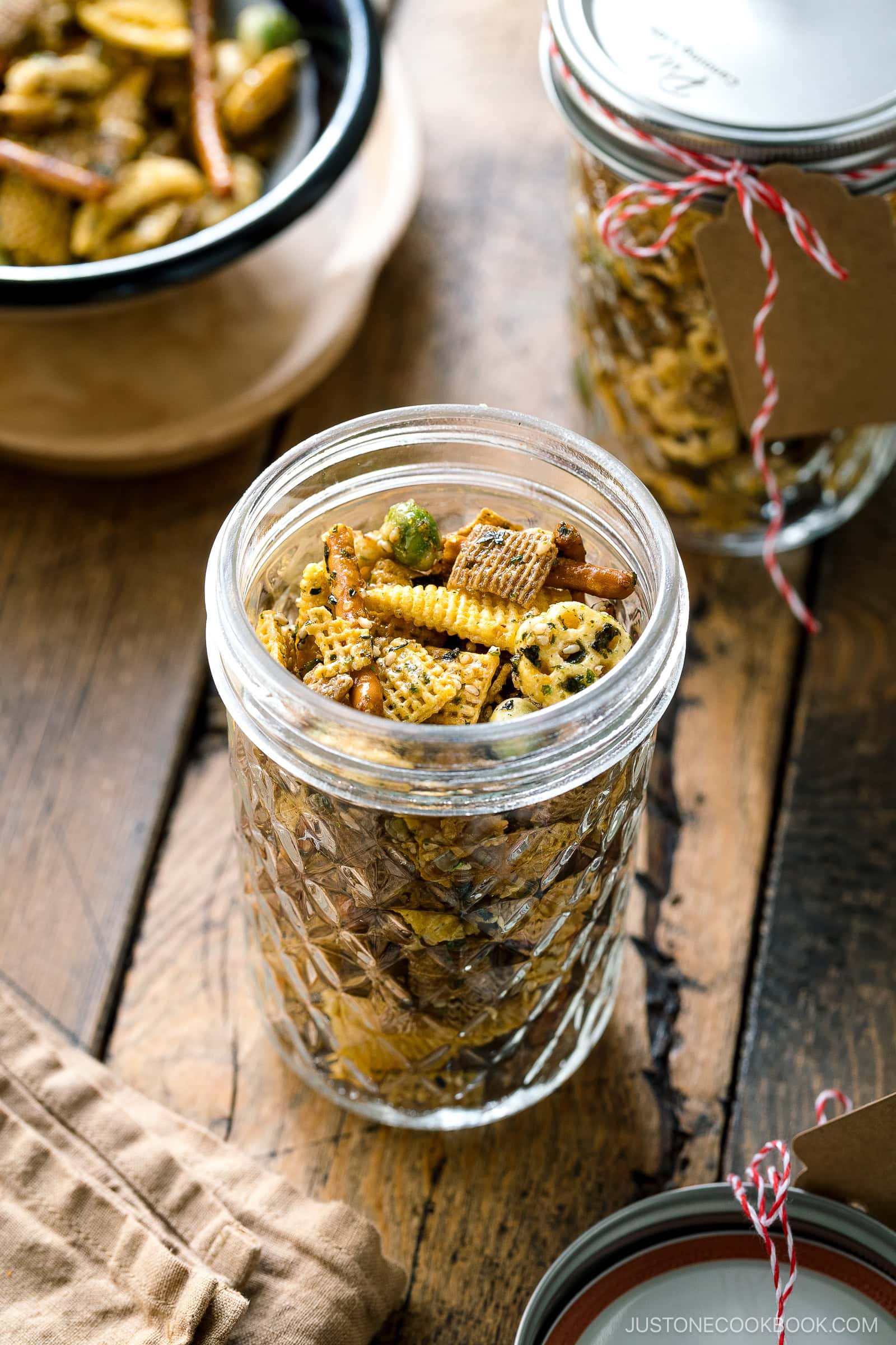 A mason jar filled with homemade snack mix, including Chex cereal, pretzels, and nuts, sits on a rustic wooden table next to another jar, a bowl of mix, and a beige napkin.