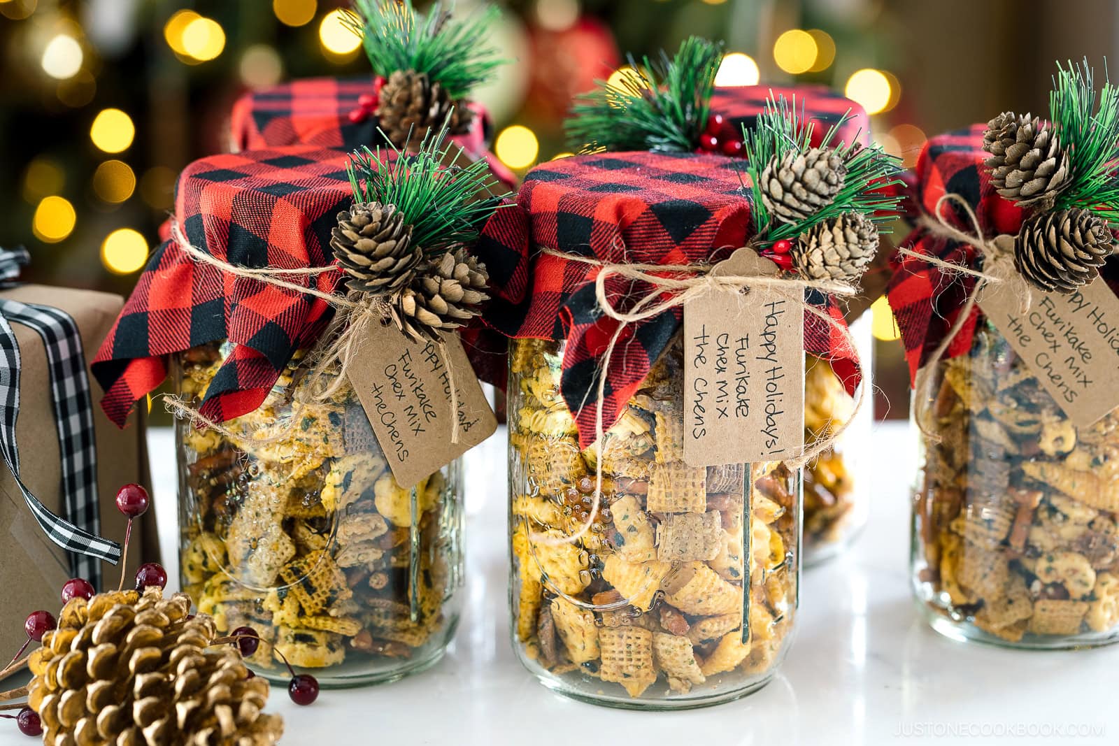 Four jars filled with snack mix are decorated with red plaid fabric, pinecones, faux pine, and holiday tags, set against a festive background with blurred Christmas lights.