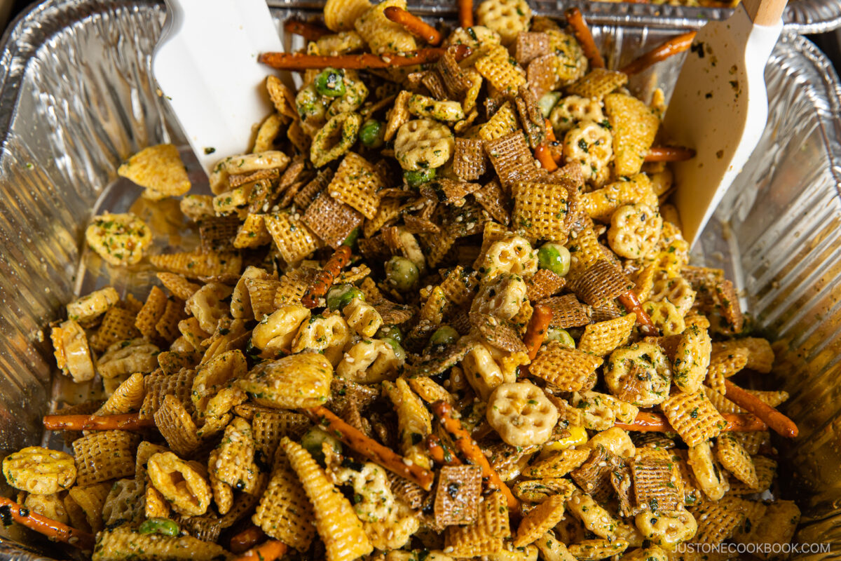 A large foil tray filled with homemade snack mix, including Chex cereal, pretzel sticks, corn chips, and other crunchy pieces coated with green seasoning. Two spatulas are visible for mixing.