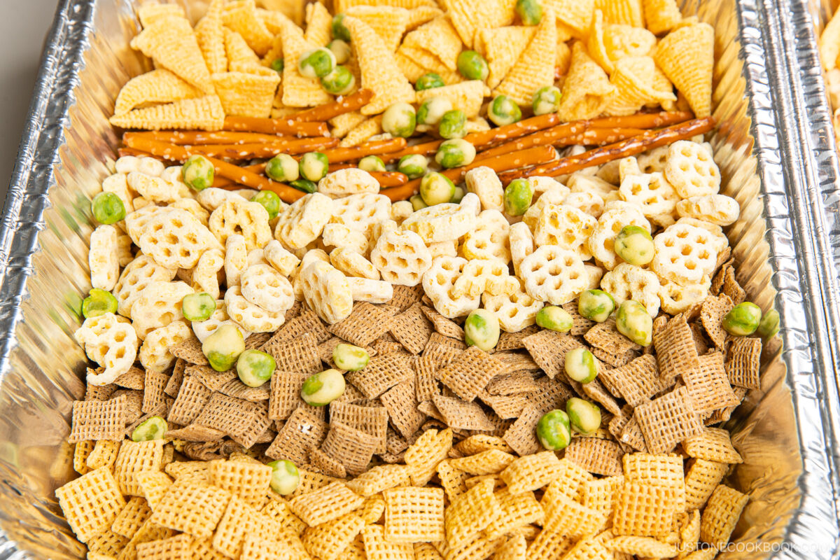 A large tray filled with an assortment of crunchy snacks, including different shapes and colors of Chex cereal, pretzel sticks, puffed snack pieces, and green wasabi peas.
