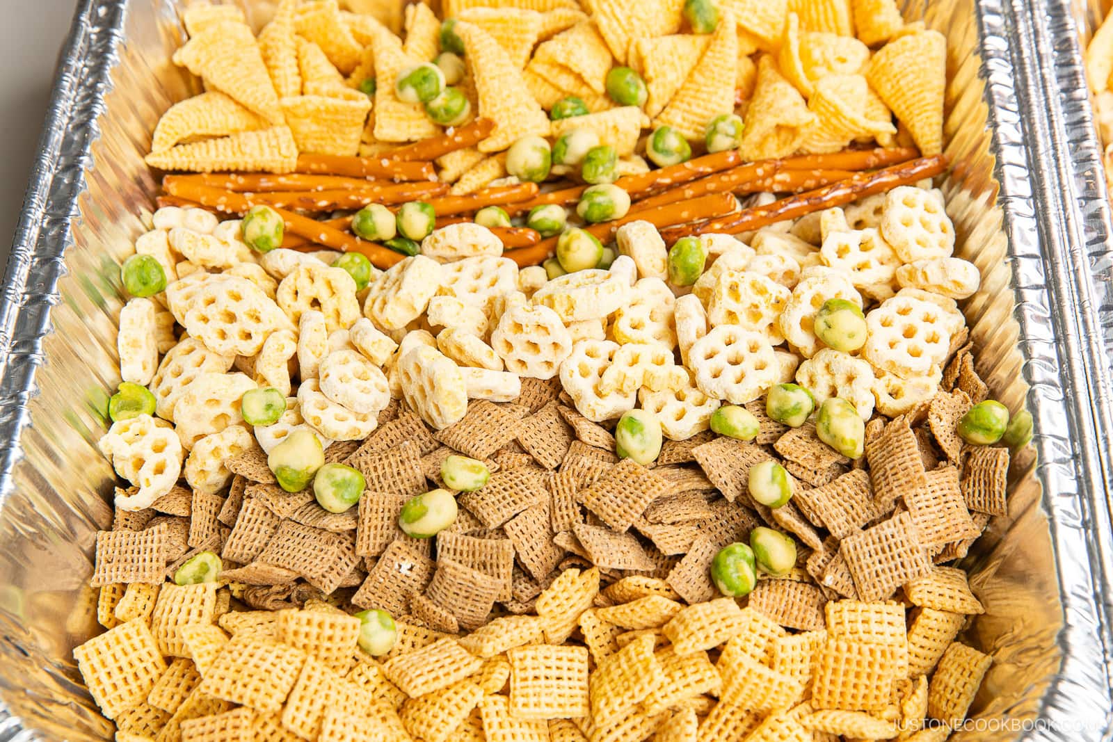 A large tray filled with an assortment of crunchy snacks, including different shapes and colors of Chex cereal, pretzel sticks, puffed snack pieces, and green wasabi peas.