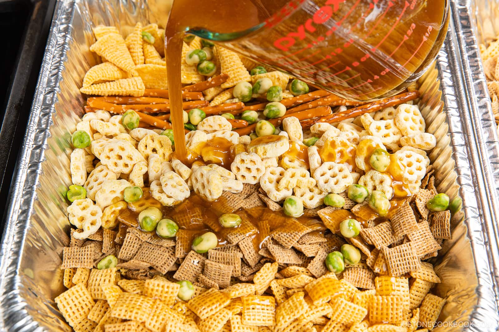 A foil tray filled with various snack mix ingredients, including cereals, pretzels, and green wasabi peas, with a measuring cup pouring brown sauce over them.