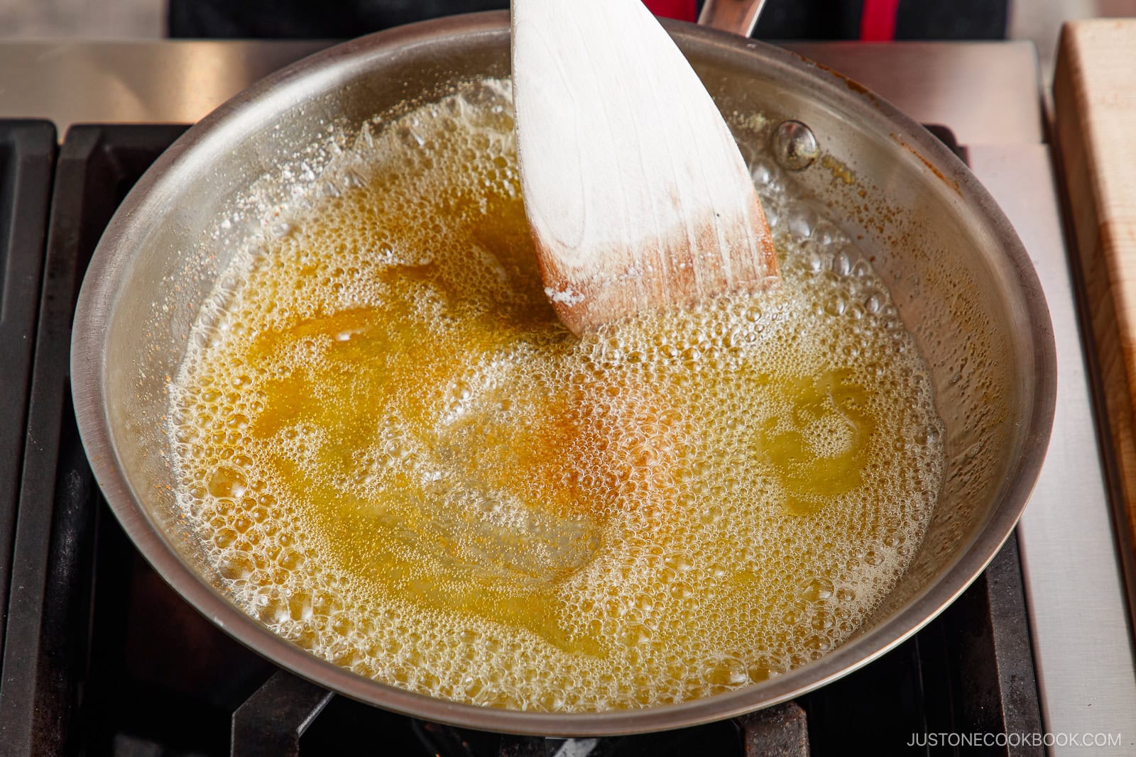A wooden spatula stirs melted butter that is bubbling and turning golden brown in a stainless steel pan on a stovetop, demonstrating how to brown butter perfectly.
