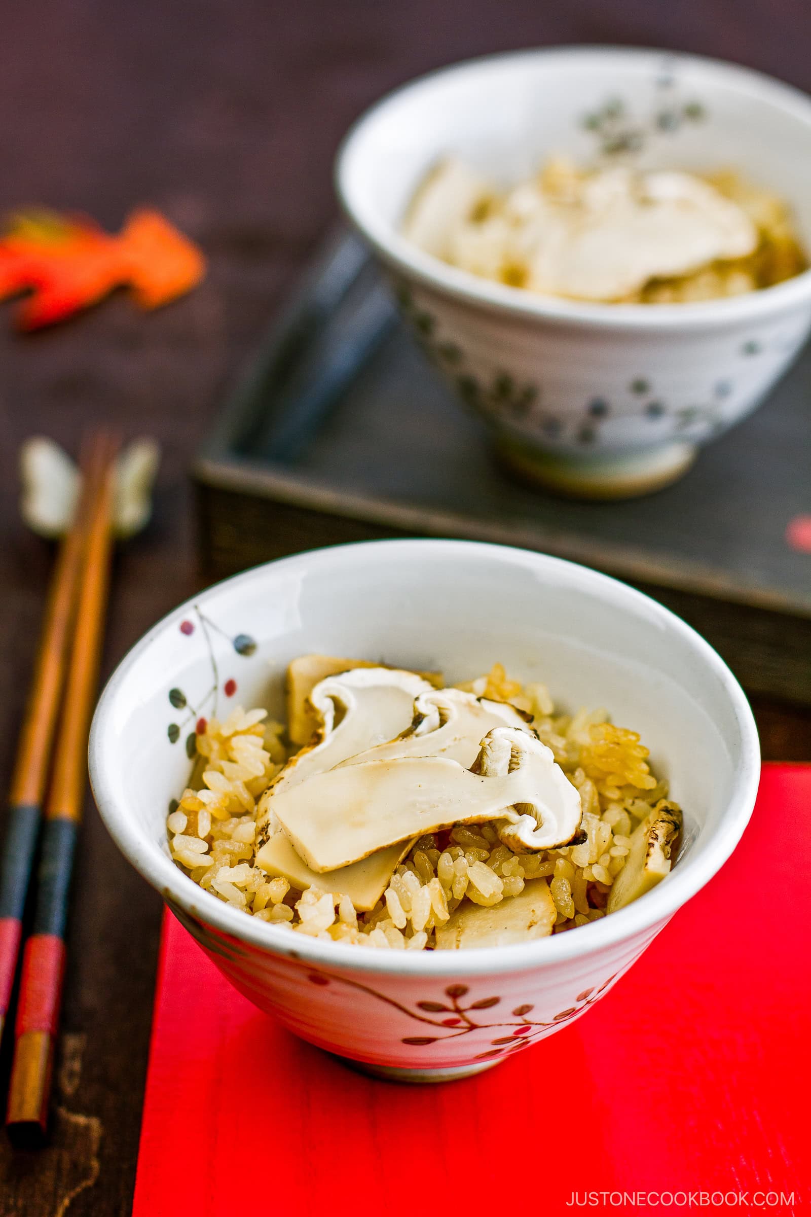 Matsutake Gohan (Wild Pine Mushroom Rice) Recipe | JustOneCookbook.com A bowl of rice topped with sliced mushrooms sits on a red mat, with another bowl and a pair of chopsticks in the background. The setting has a warm, rustic atmosphere.