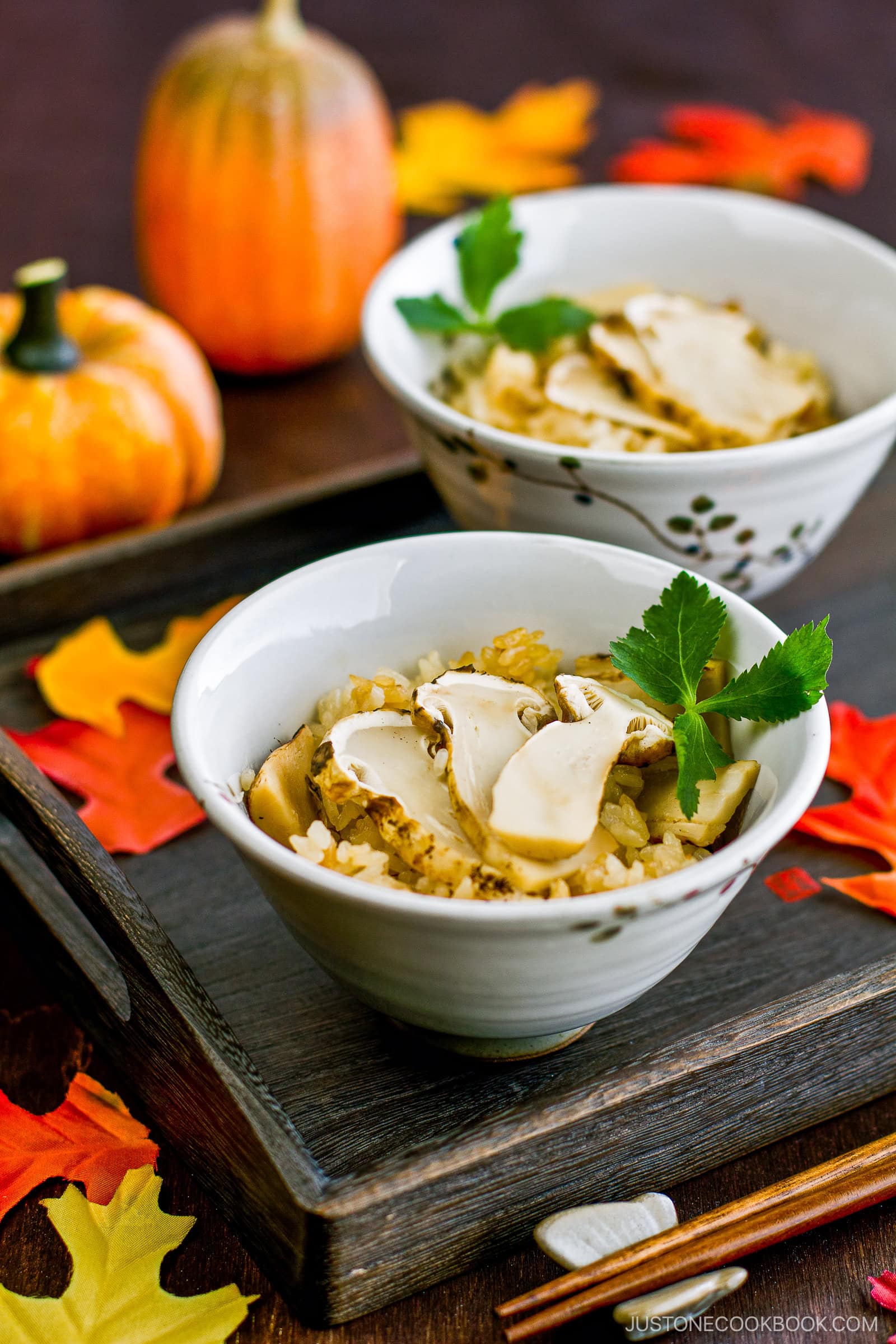 Matsutake Gohan II Two bowls of rice topped with sliced mushrooms and garnished with green herbs are served on a wooden tray, surrounded by autumn leaves and small pumpkins. Chopsticks rest nearby, creating a cozy fall atmosphere.