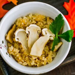 A white bowl filled with seasoned rice and sliced mushrooms, garnished with a green leaf. The bowl sits on a tray decorated with artificial autumn leaves.