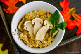 A white bowl filled with seasoned rice and sliced mushrooms, garnished with a green leaf. The bowl sits on a tray decorated with artificial autumn leaves.
