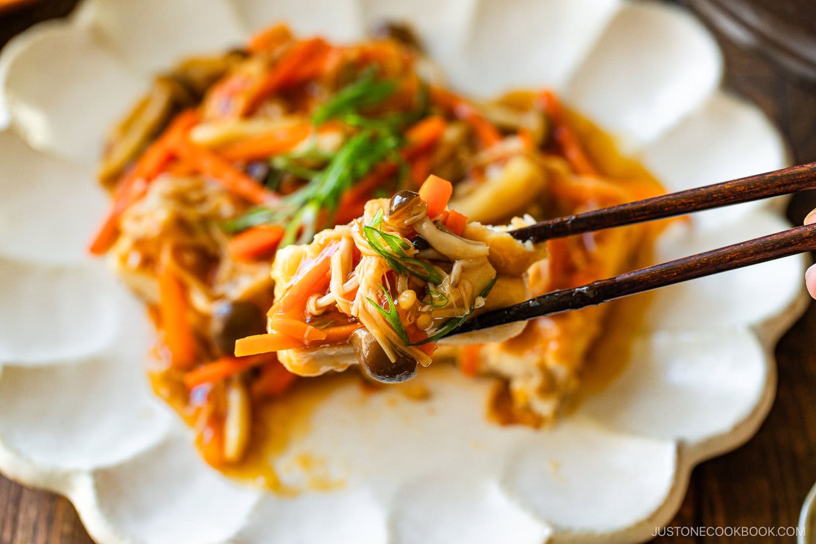 Chopsticks holding a bite of colorful stir-fried vegetables and mushrooms over a white, scalloped plate with more of the dish in the background.