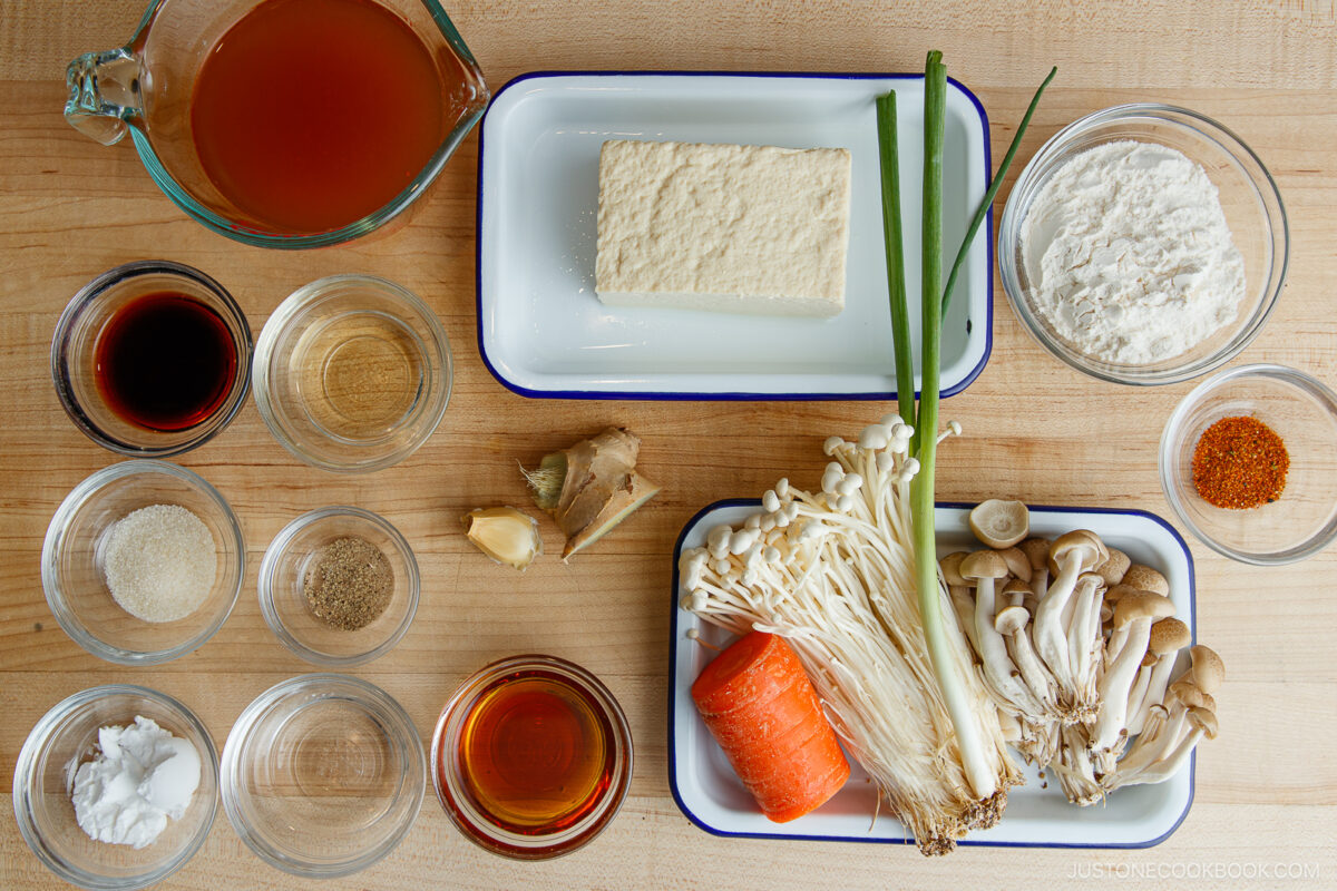 Top-down view of various ingredients on a wooden surface, including tofu, mushrooms, carrot, green onion, flour, soy sauce, vinegar, broth, ginger, sugar, pepper, and seasonings in small bowls.