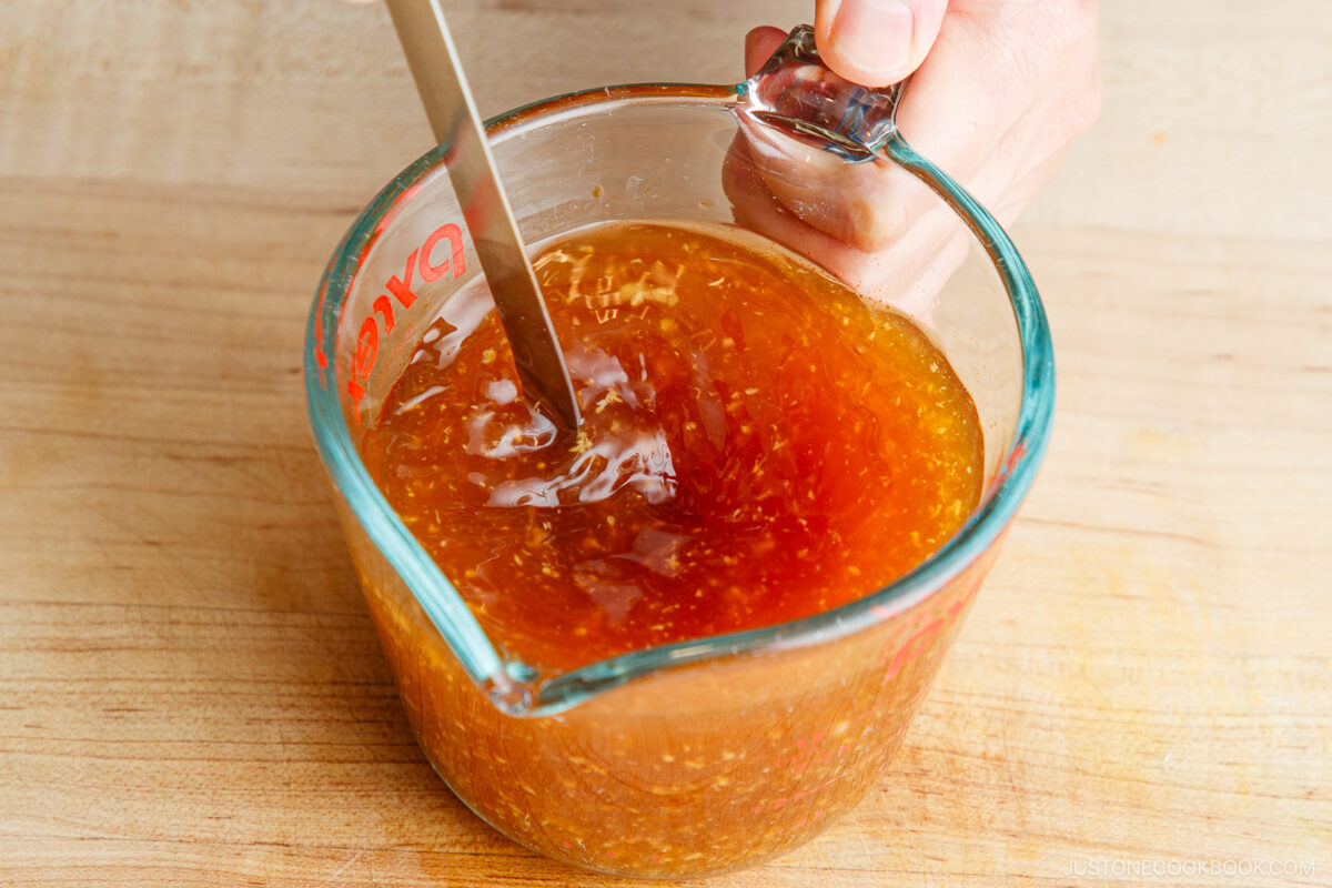 A hand holds a glass measuring cup filled with orange-brown sauce, stirring it with a metal spoon on a wooden surface.