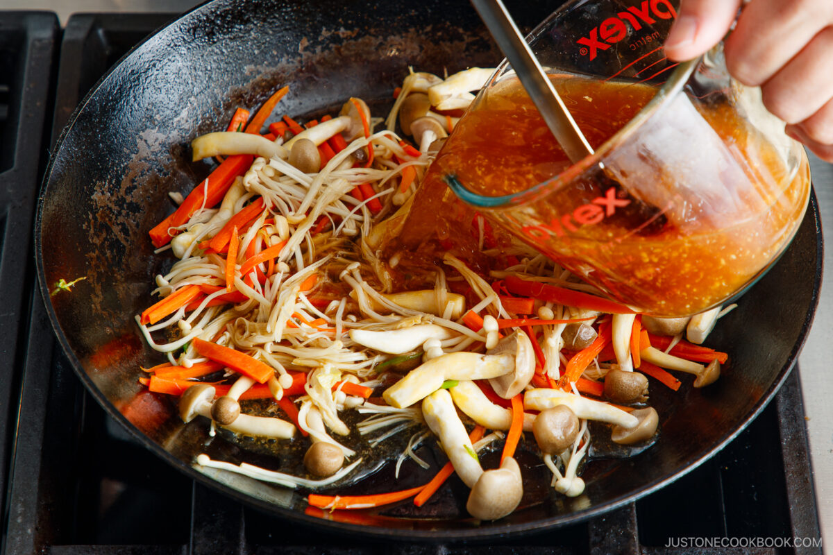 A hand pours sauce from a measuring cup into a pan filled with sautéing mushrooms and julienned carrots on a stovetop.