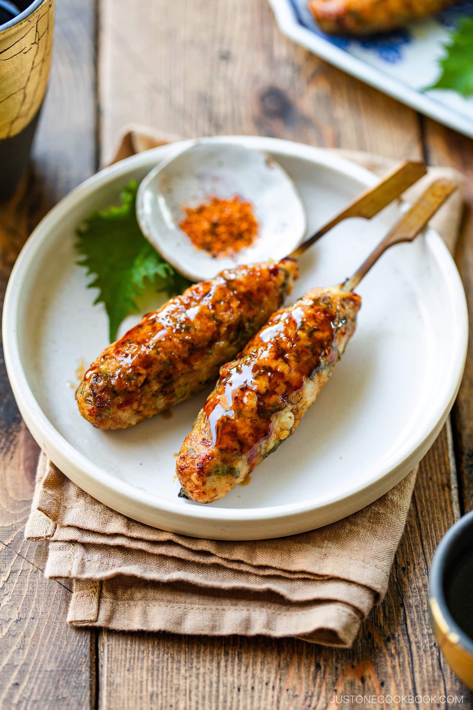 Two skewers of glazed Japanese chicken tsukune sit on a white plate, garnished with sauce. A small dish with red spice and a shiso leaf are beside them, all set on a folded cloth atop a rustic wooden table.