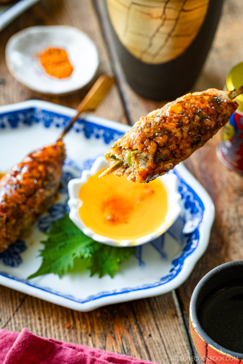 A close-up of grilled meat skewers, glazed with sauce, served on a blue and white plate with a leaf garnish and a small bowl of dipping sauce. A cup and spice dish are in the background on a wooden table.