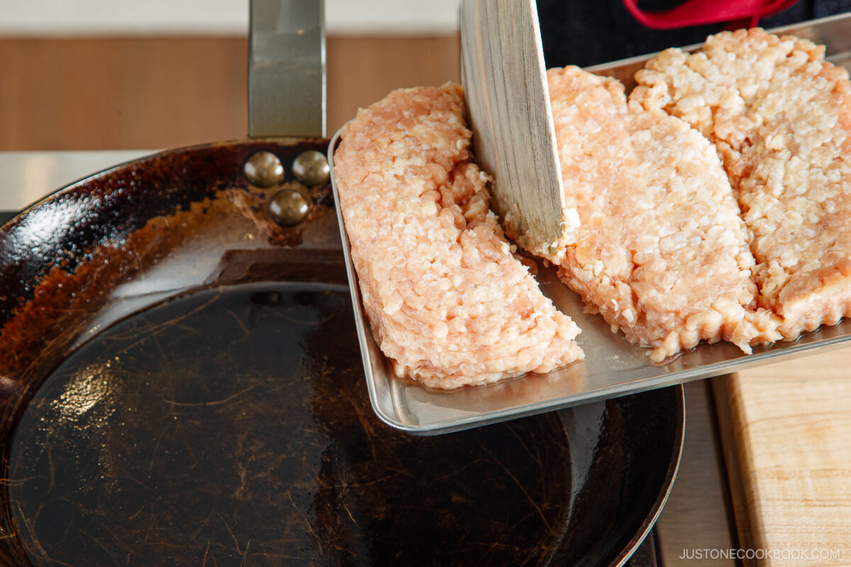 Raw ground meat is being transferred from a metal tray into a heated frying pan, ready to be cooked.