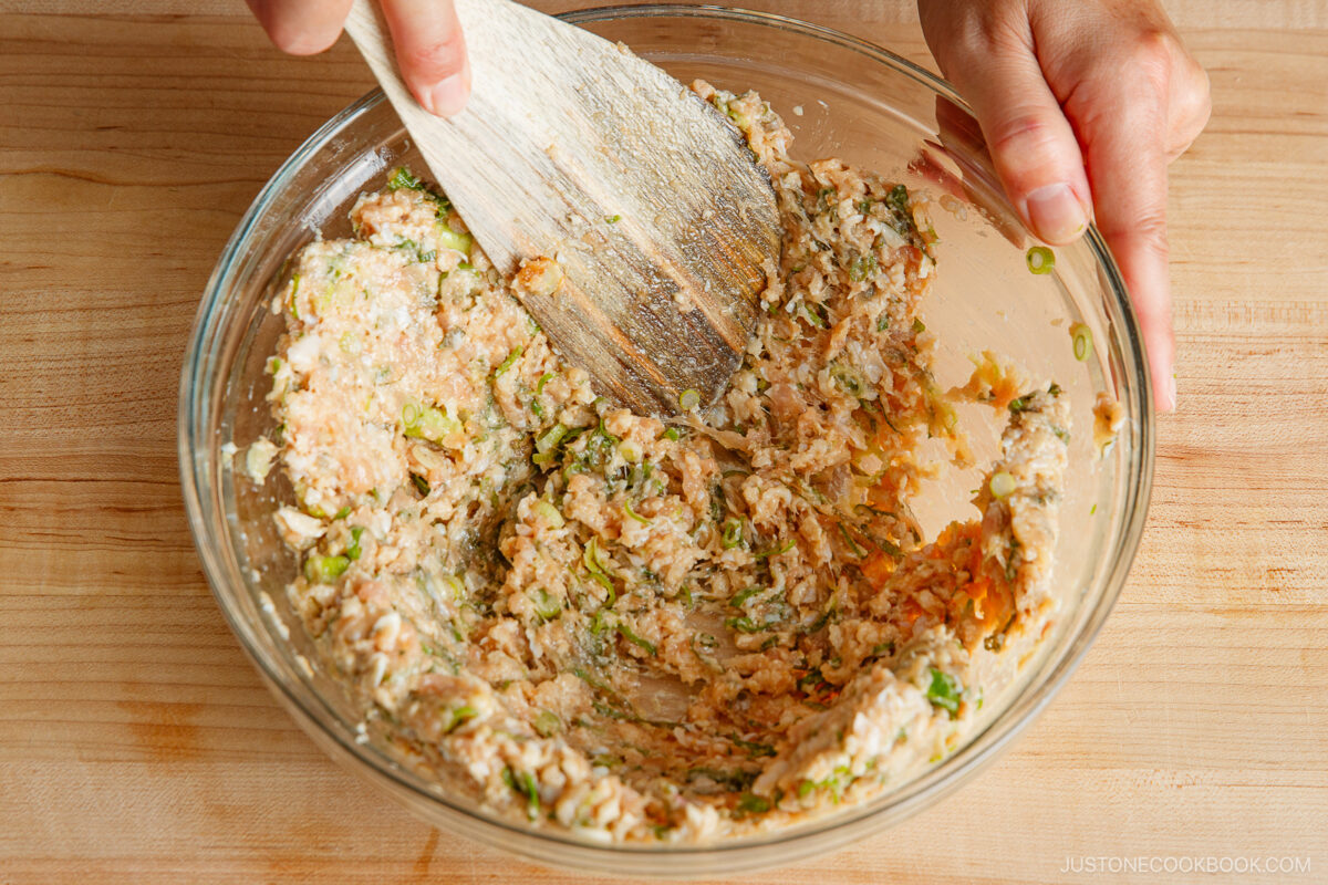 Hands mixing ground meat, chopped vegetables, and seasonings in a glass bowl using a wooden spatula on a wooden surface.