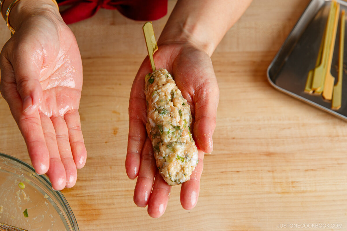 A person shapes seasoned ground meat onto a gold skewer with their hands over a wooden surface, preparing kebabs. A metal tray with more skewers is visible in the background.