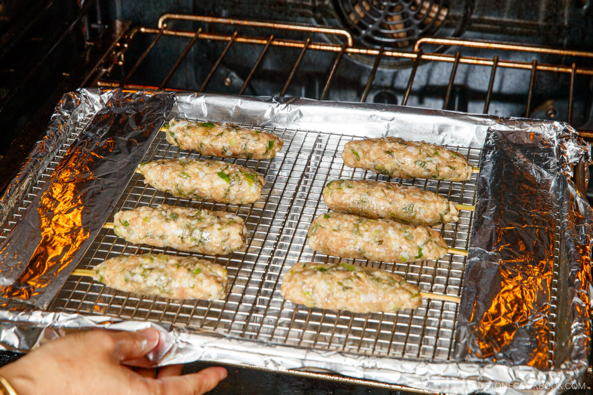 Raw kebabs on skewers are arranged on a wire rack over a foil-lined baking sheet, being placed into an oven for cooking. A hand is holding the tray, and the oven racks are visible inside.