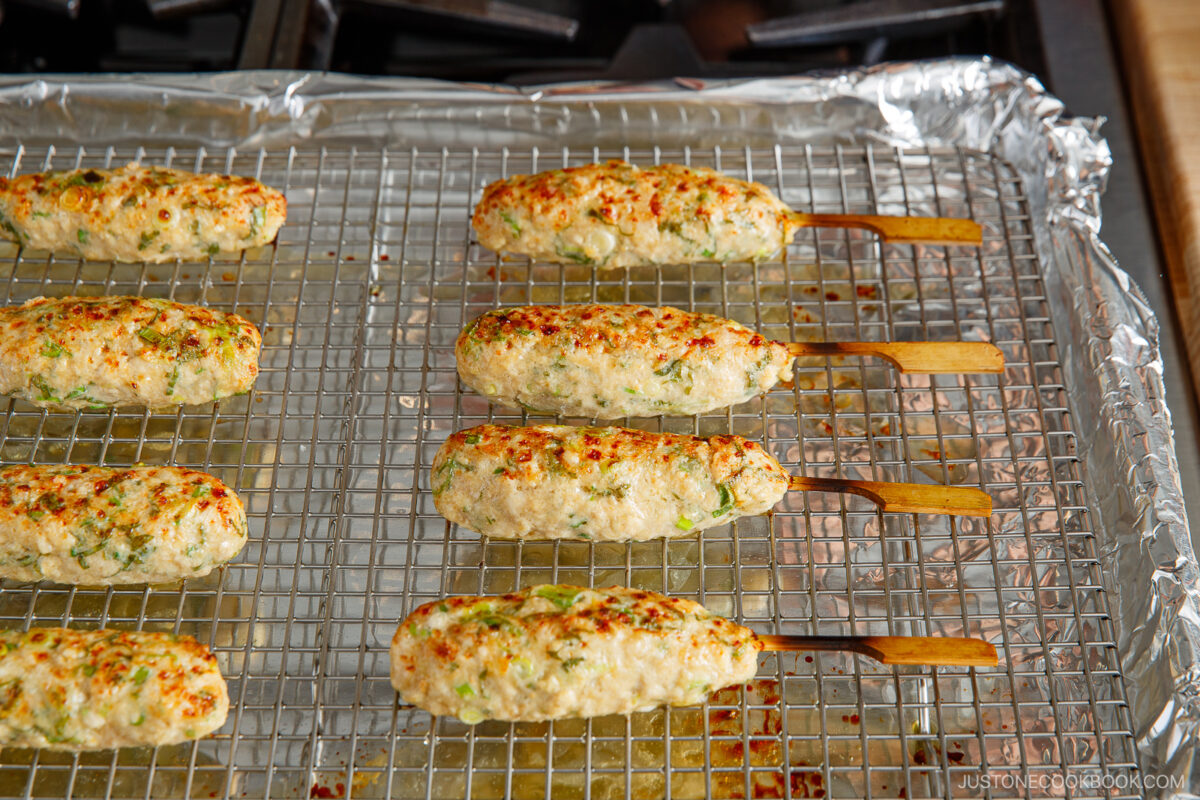 Seven grilled chicken tsukune skewers rest on a wire rack over a foil-lined baking sheet. The skewers are golden brown and speckled with green herbs, set on a stovetop.