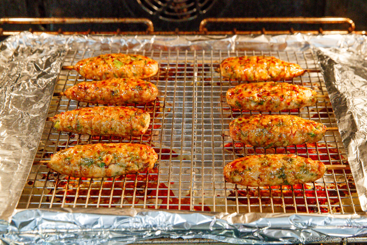 Chicken kebabs are baking in an oven, arranged in two rows on a wire rack over a foil-lined baking sheet. The kebabs are golden brown and speckled with herbs and seasonings.