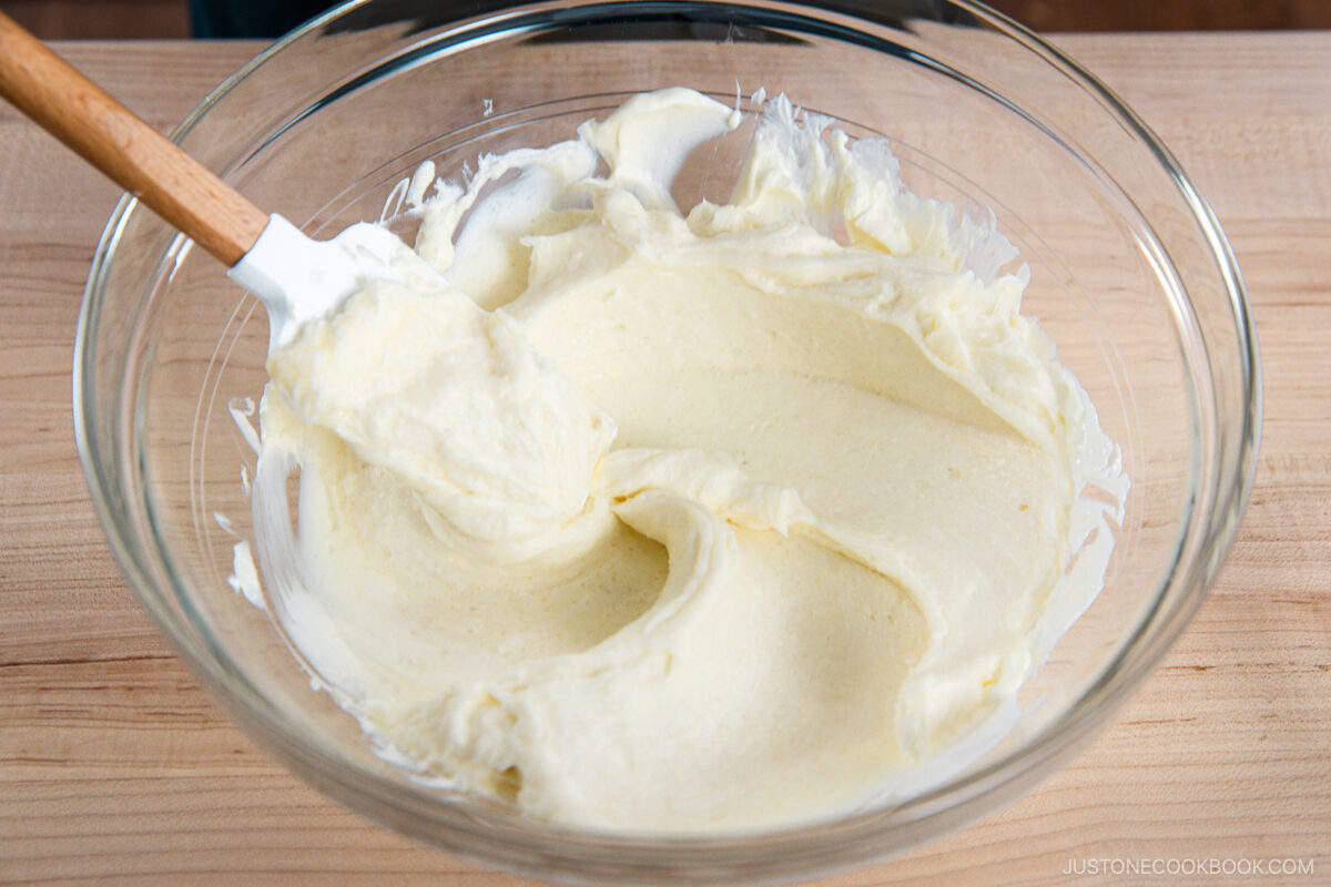 A glass mixing bowl filled with creamy cheesecake batter being stirred with a spatula, placed on a wooden surface.