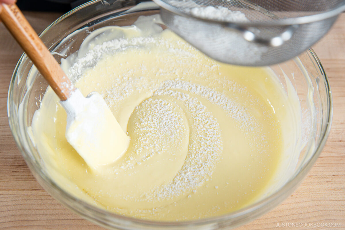A bowl of smooth yellow cake batter being mixed with a spatula as powdered ingredients are being sifted in from a fine mesh strainer above.