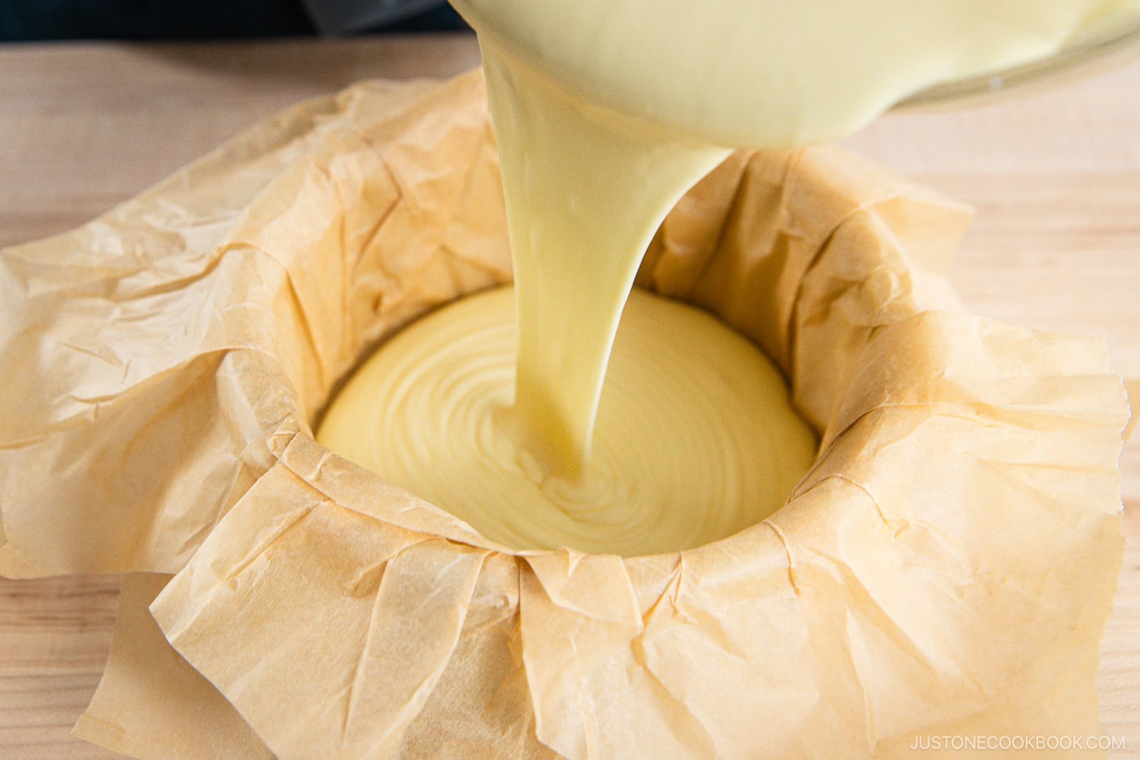 Pale yellow batter is being poured into a round cake pan lined with crumpled parchment paper, resting on a light wooden surface.
