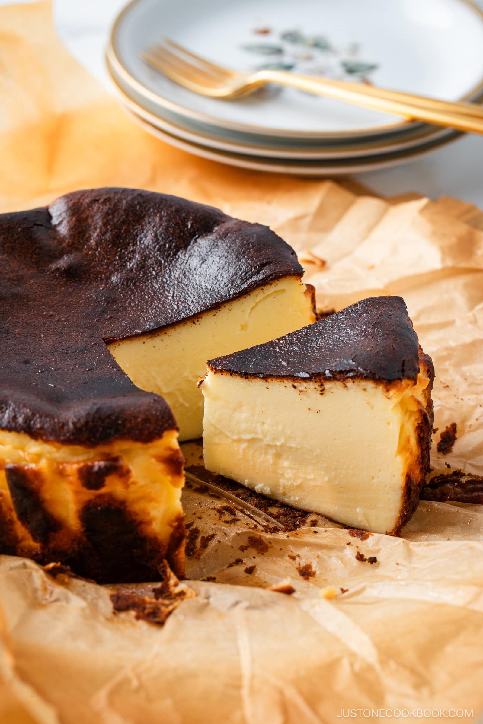 A sliced Basque burnt cheesecake sits on parchment paper, with its creamy interior and caramelized top visible. Plates and a fork are stacked in the background.