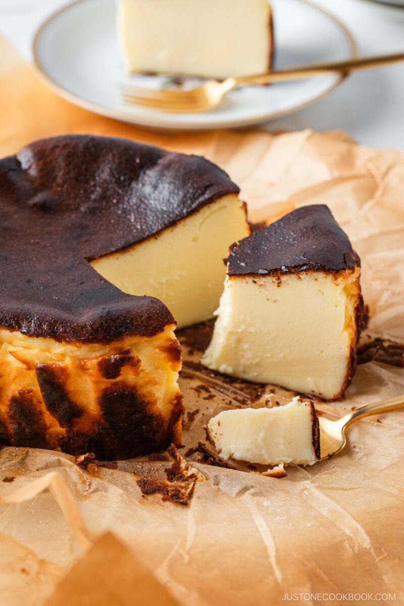 A sliced Basque burnt cheesecake on parchment paper, with a golden brown, slightly burnt top and creamy interior. A fork and a serving plate with a cheesecake slice are in the background.
