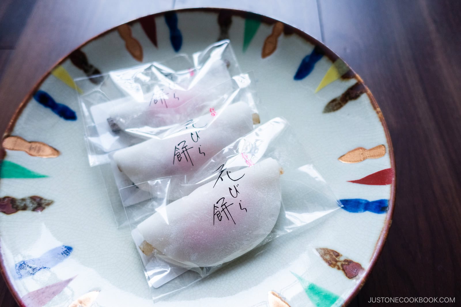 A decorative plate holds several individually wrapped pieces of Japanese confectionery, likely mochi, each in clear plastic packaging with Japanese writing on it. The plate rests on a dark wooden surface.