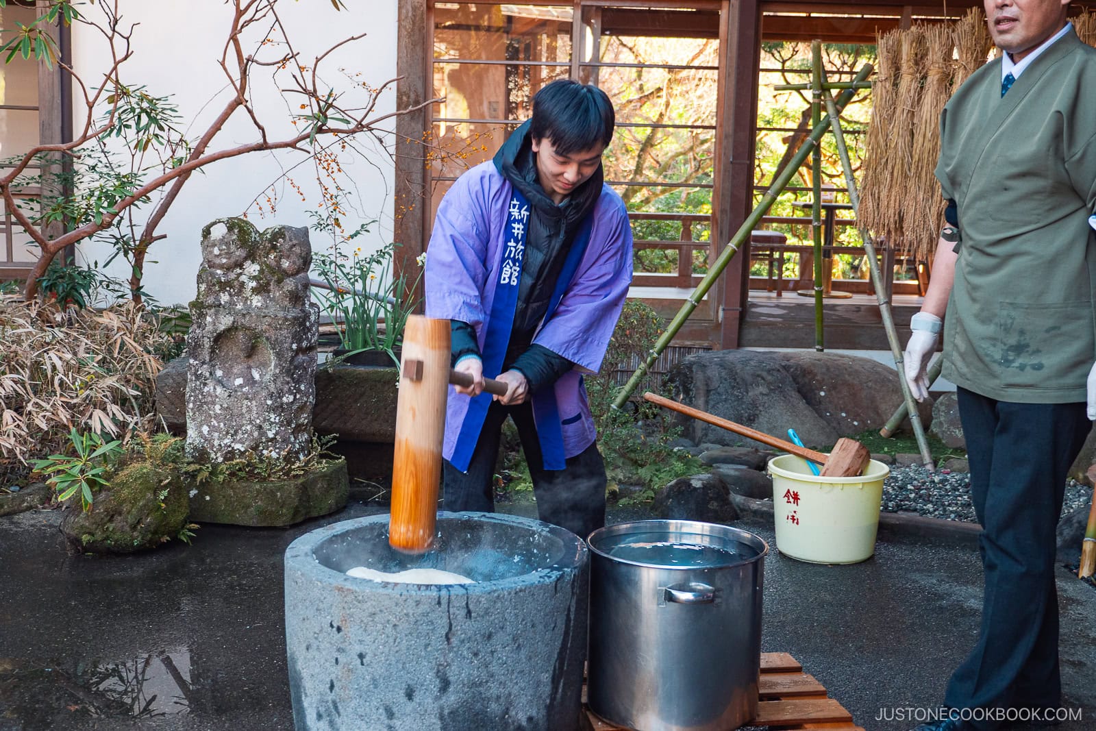 A man in traditional Japanese clothing pounds steamed rice with a wooden mallet in an outdoor stone mortar for mochi making, while another person stands nearby. Pots and water are placed next to the mortar.