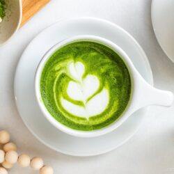 A top view of a white cup filled with matcha latte, featuring latte art on the frothy green surface. Surrounding items include a whisk, beads, a cloth napkin, and a bowl of matcha powder.