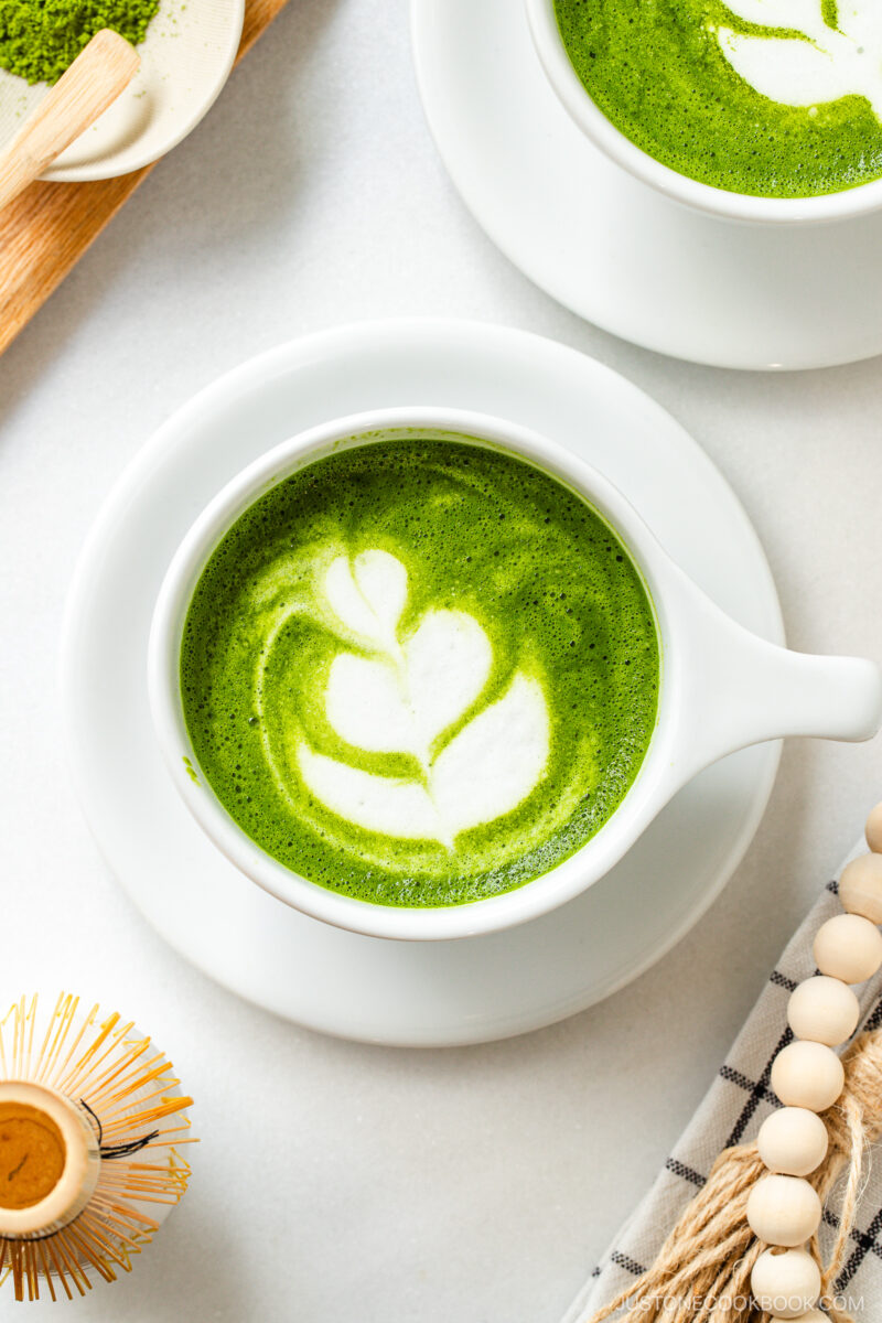 A cup of matcha latte with white leaf-shaped latte art on top, served in a white cup and saucer, with a bamboo whisk, cloth napkin, and other cups nearby on a light-colored surface.