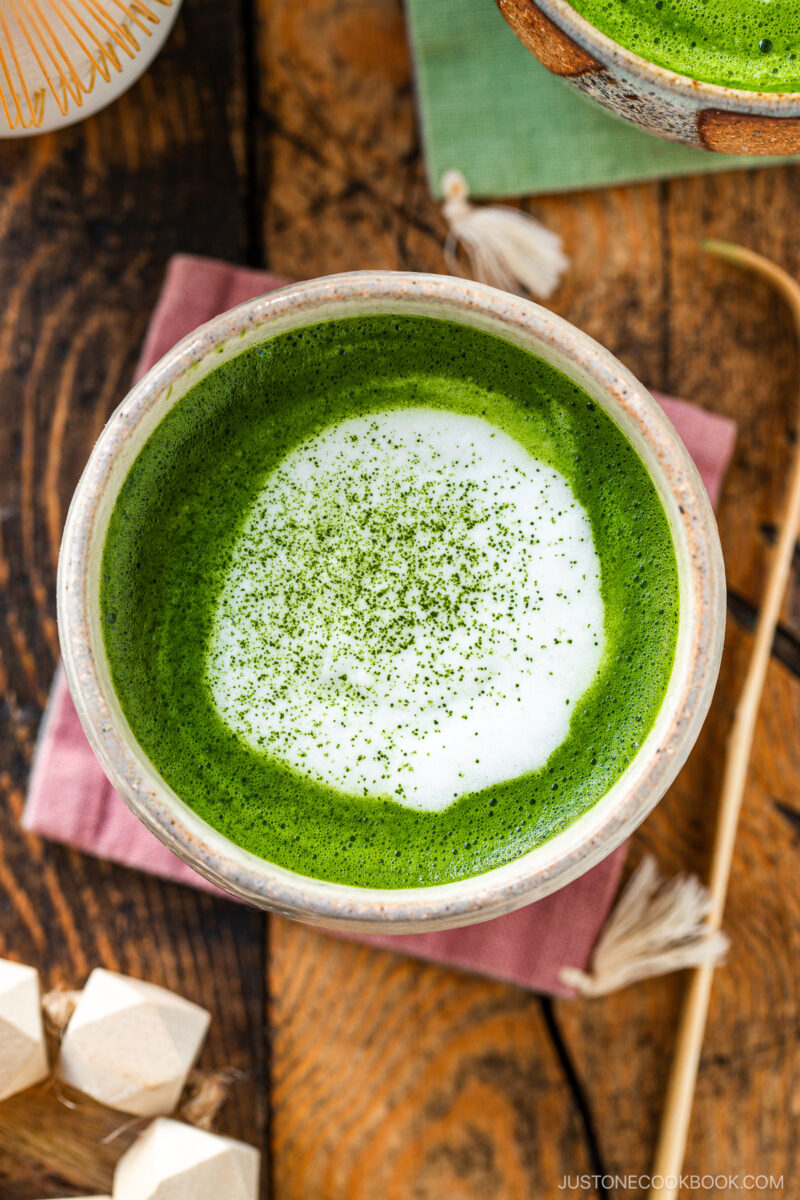 A ceramic bowl filled with vibrant green matcha latte topped with creamy white foam and a sprinkle of matcha powder, placed on a pink napkin on a wooden table.