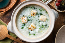 A ceramic pot filled with rice porridge topped with grilled tofu cubes and chopped greens sits on a wooden table, next to a wooden spoon and various dishes.