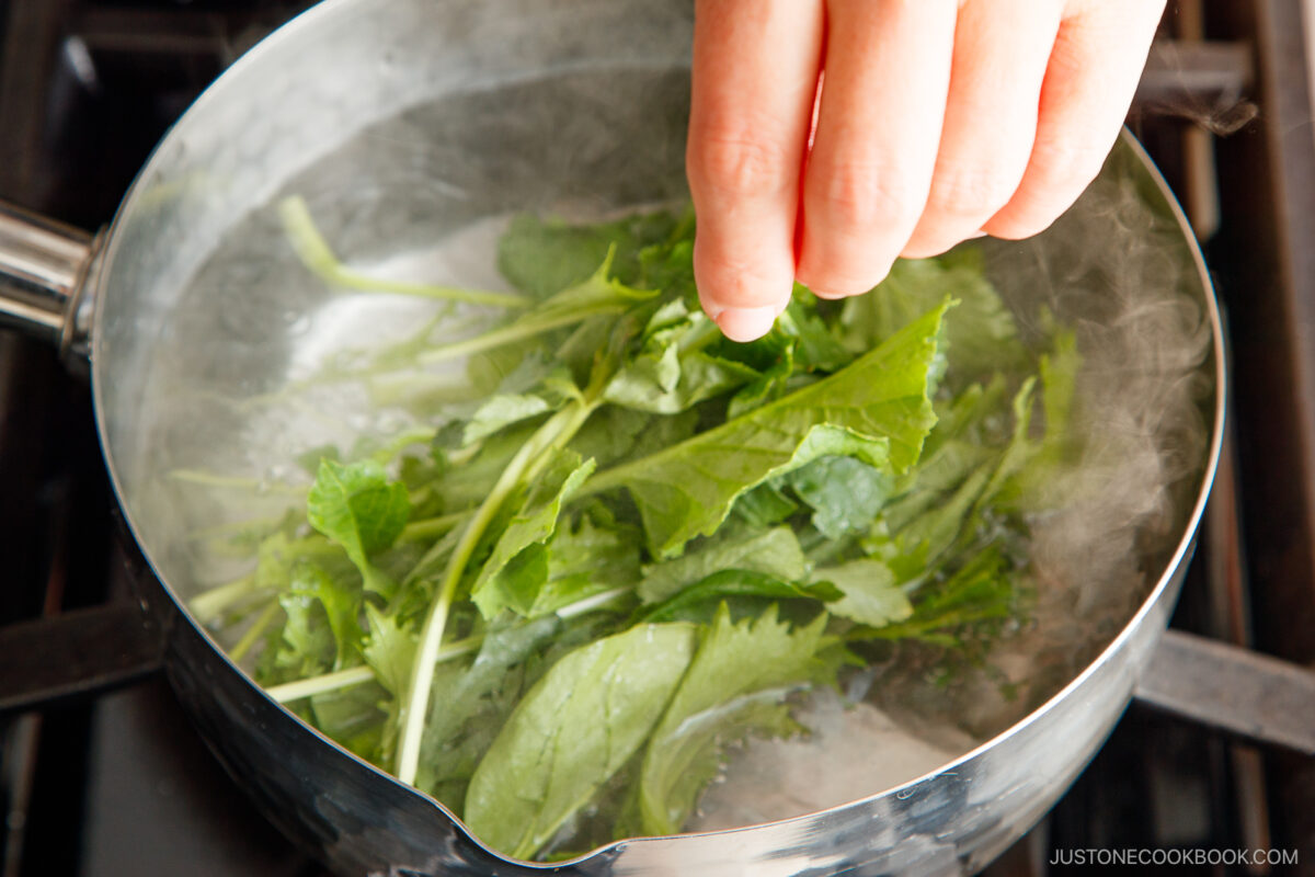A hand adds fresh leafy greens into a pot of boiling water on a stovetop, with steam rising from the pot.