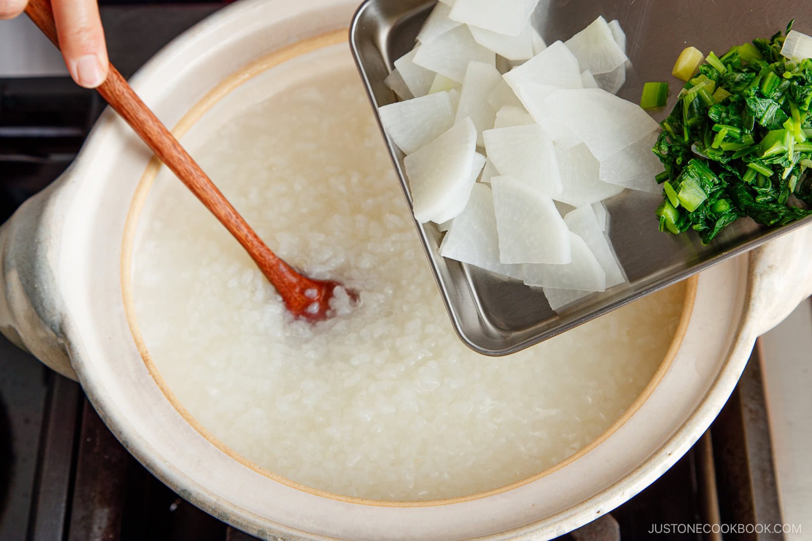 A hand stirs a pot of rice porridge with a wooden spoon while adding sliced white radish and chopped green vegetables from a metal tray.