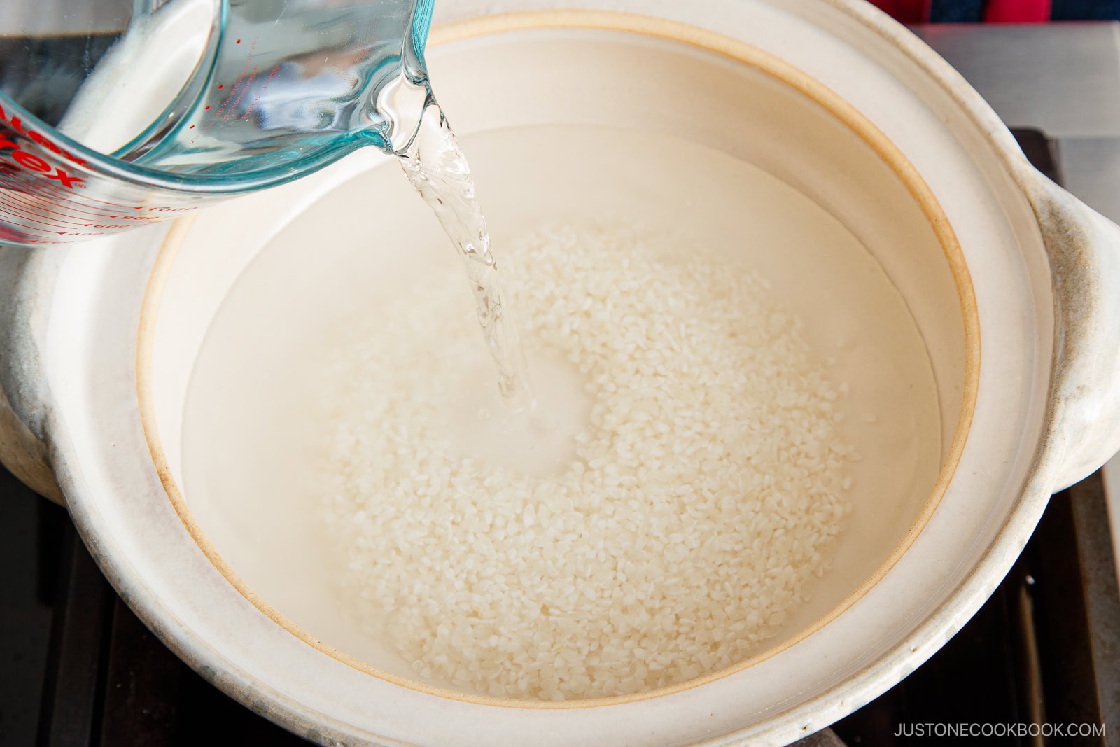 A hand pours water from a glass measuring cup into a white pot filled with uncooked white rice, preparing to cook the rice on a stovetop.