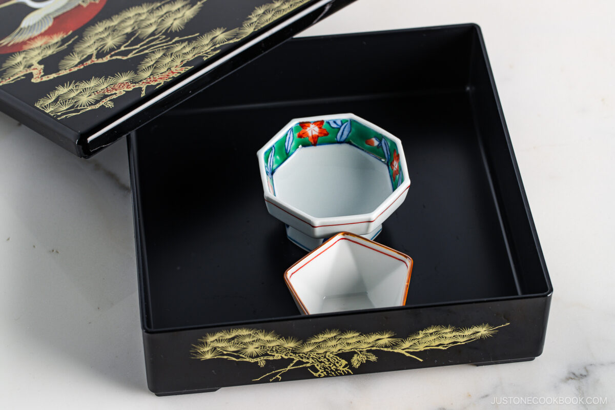 A black lacquered Japanese bento box with gold pine tree designs holds two small, empty porcelain bowls—one octagonal with colorful patterns and one hexagonal with a red rim. The box lid is partially open.