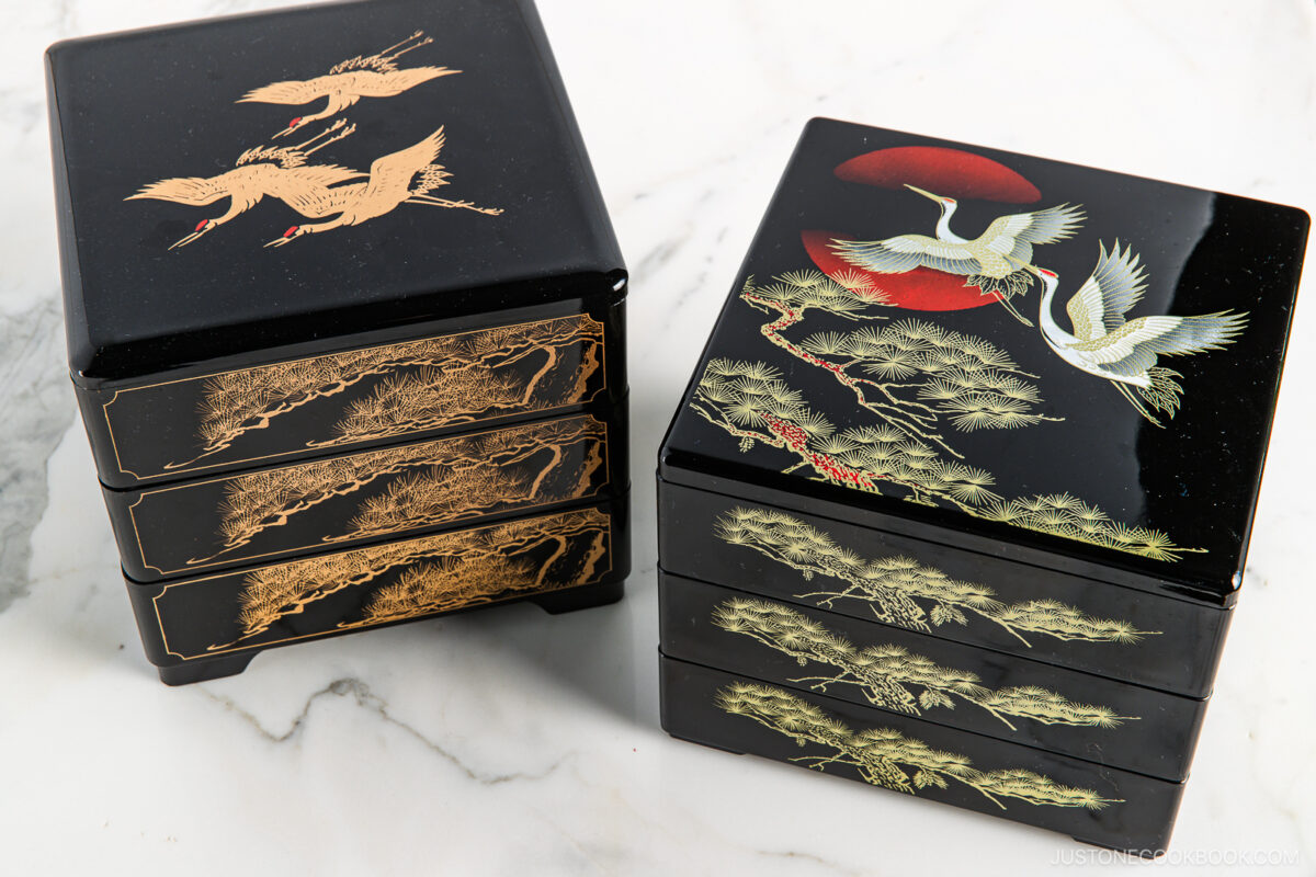 Two black Japanese bento boxes with three tiers each, decorated with gold and colored crane and pine tree designs, placed on a white marble surface.