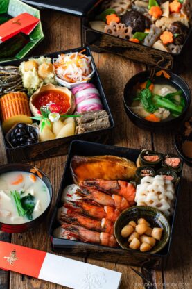 A colorful arrangement of traditional Japanese New Year’s foods, including shrimp, fish, vegetables, and assorted delicacies in lacquered boxes, with bowls of soup on a wooden table.