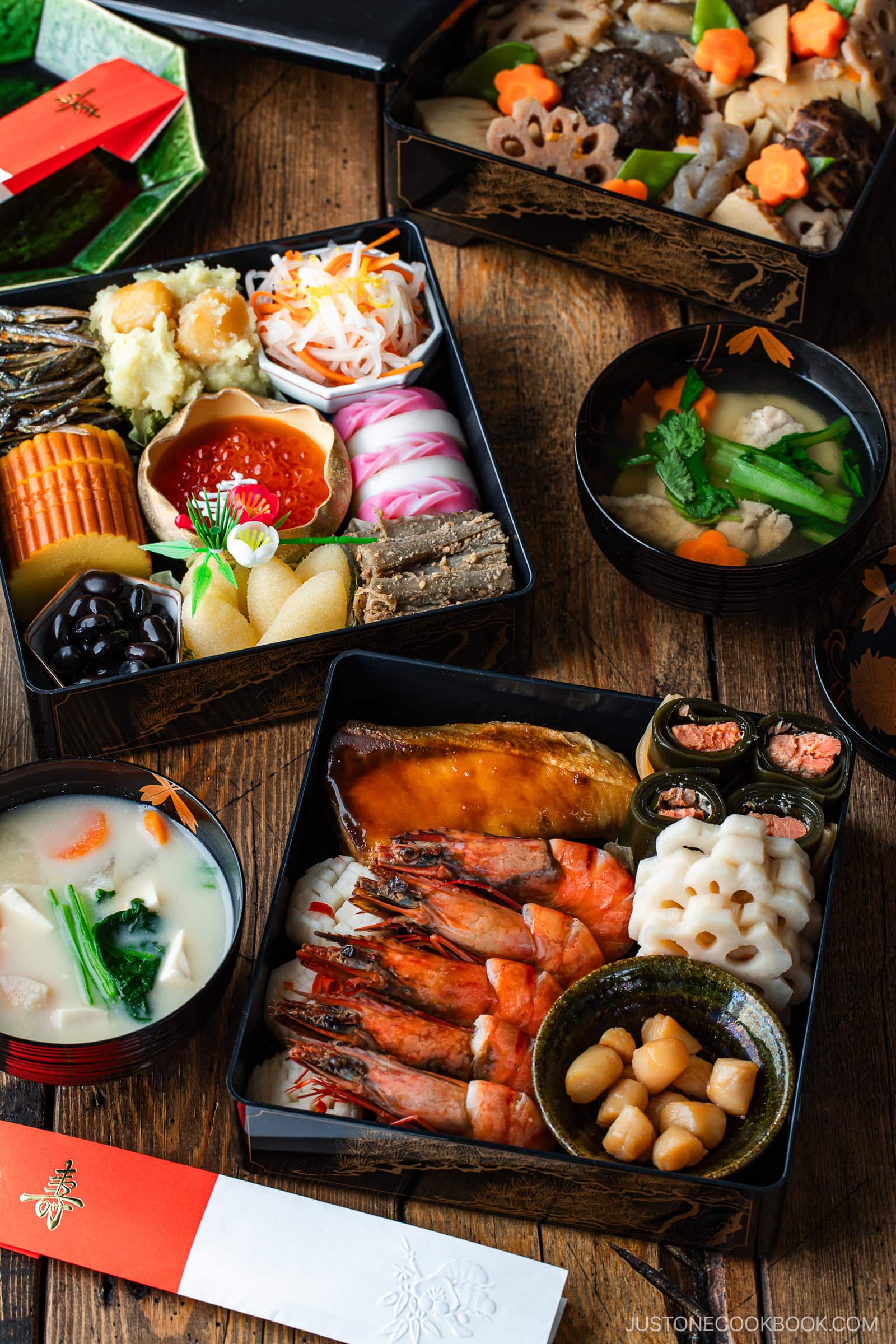 A colorful arrangement of traditional Japanese New Year’s foods, including shrimp, fish, vegetables, and assorted delicacies in lacquered boxes, with bowls of soup on a wooden table.