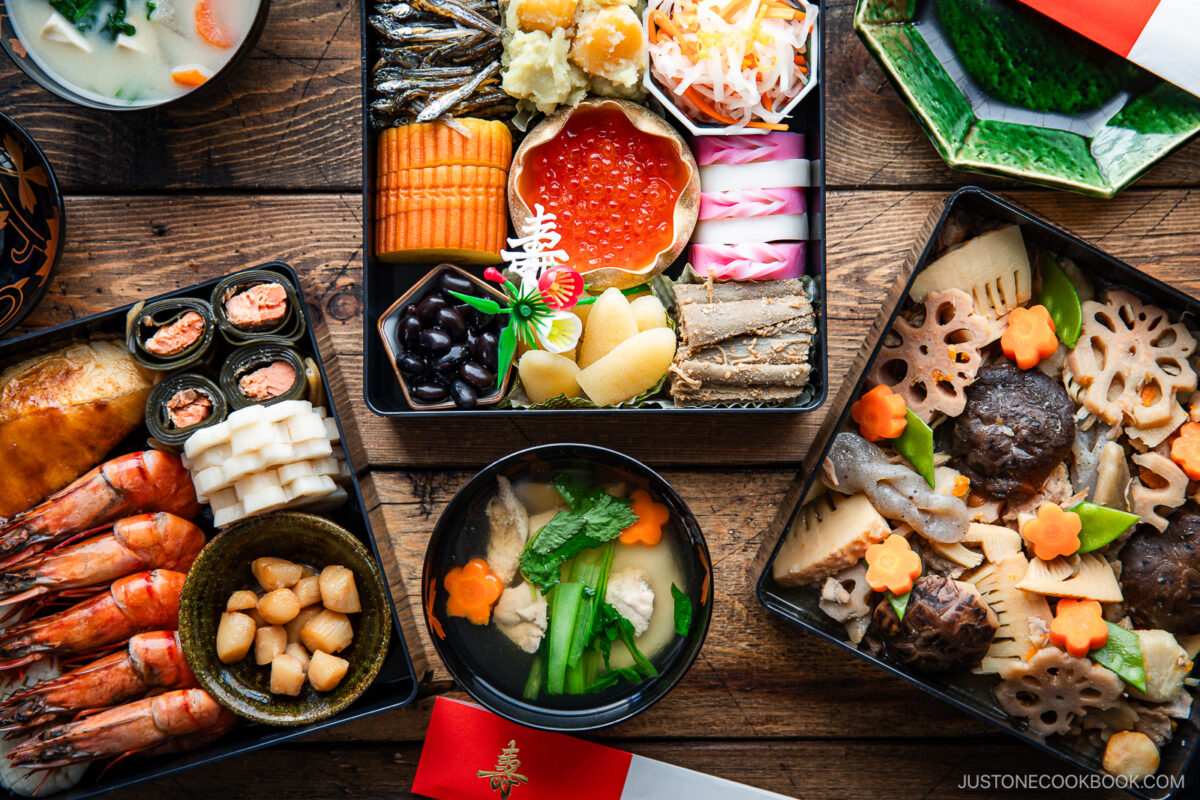 An assortment of traditional Japanese New Year foods, including colorful seafood, vegetables, and sweets arranged in lacquered boxes and bowls on a wooden table.
