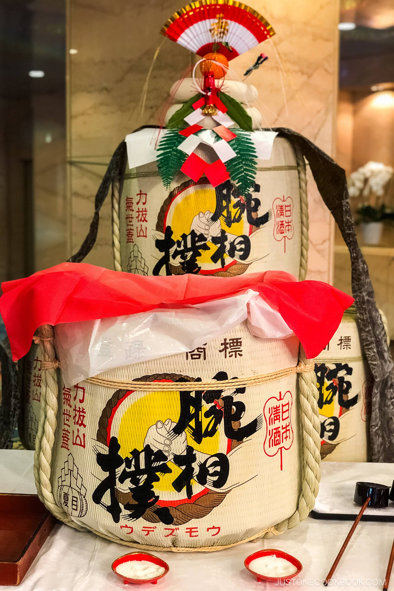 Large decorative sake barrels wrapped in straw, adorned with Japanese writing and symbols. The front barrel is open and covered with a red cloth, and festive ornaments are displayed on top. The background shows a marble wall.
