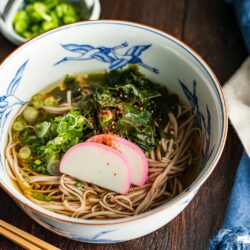 A bowl of soba noodle soup topped with sliced fish cake, seaweed, chopped green onions, and seasoning, with chopsticks and a blue napkin beside it on a wooden table.