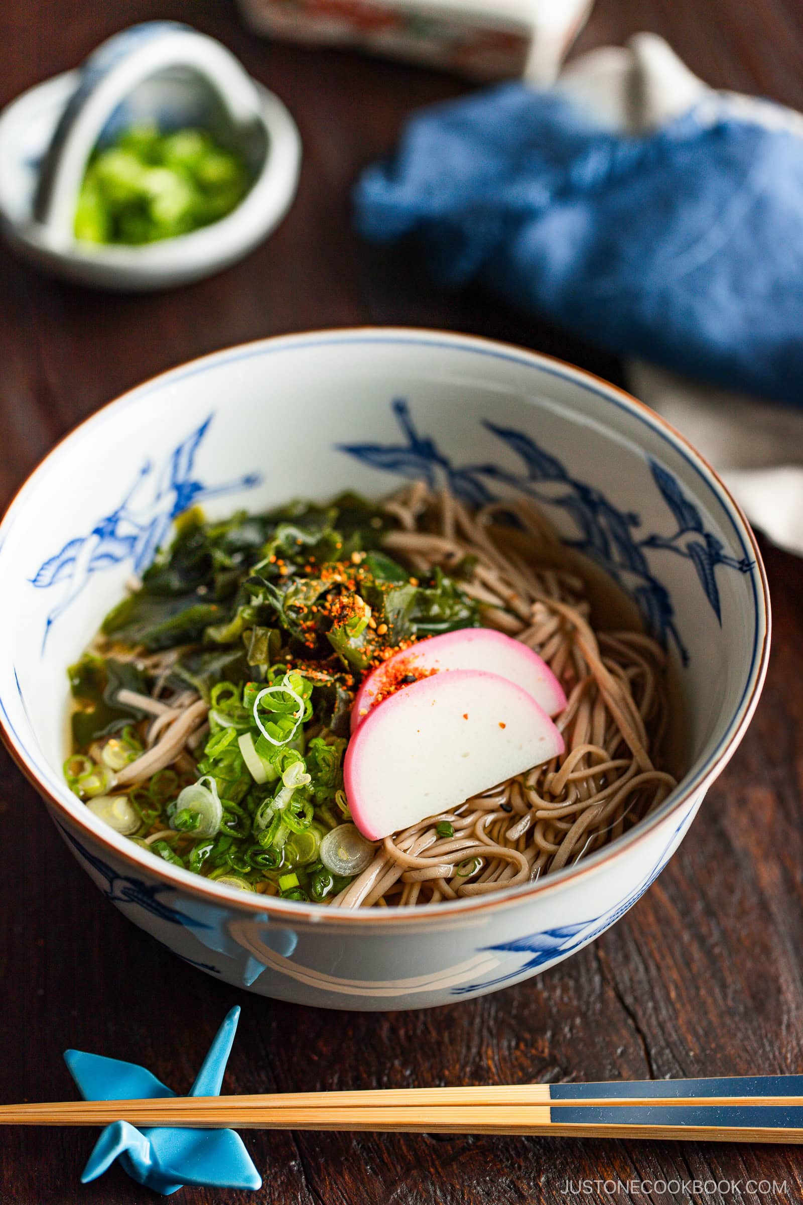 A bowl of soba noodles in broth, topped with sliced fish cake, green onions, and seaweed, sits on a wooden table with chopsticks and a small dish of chopped scallions nearby.