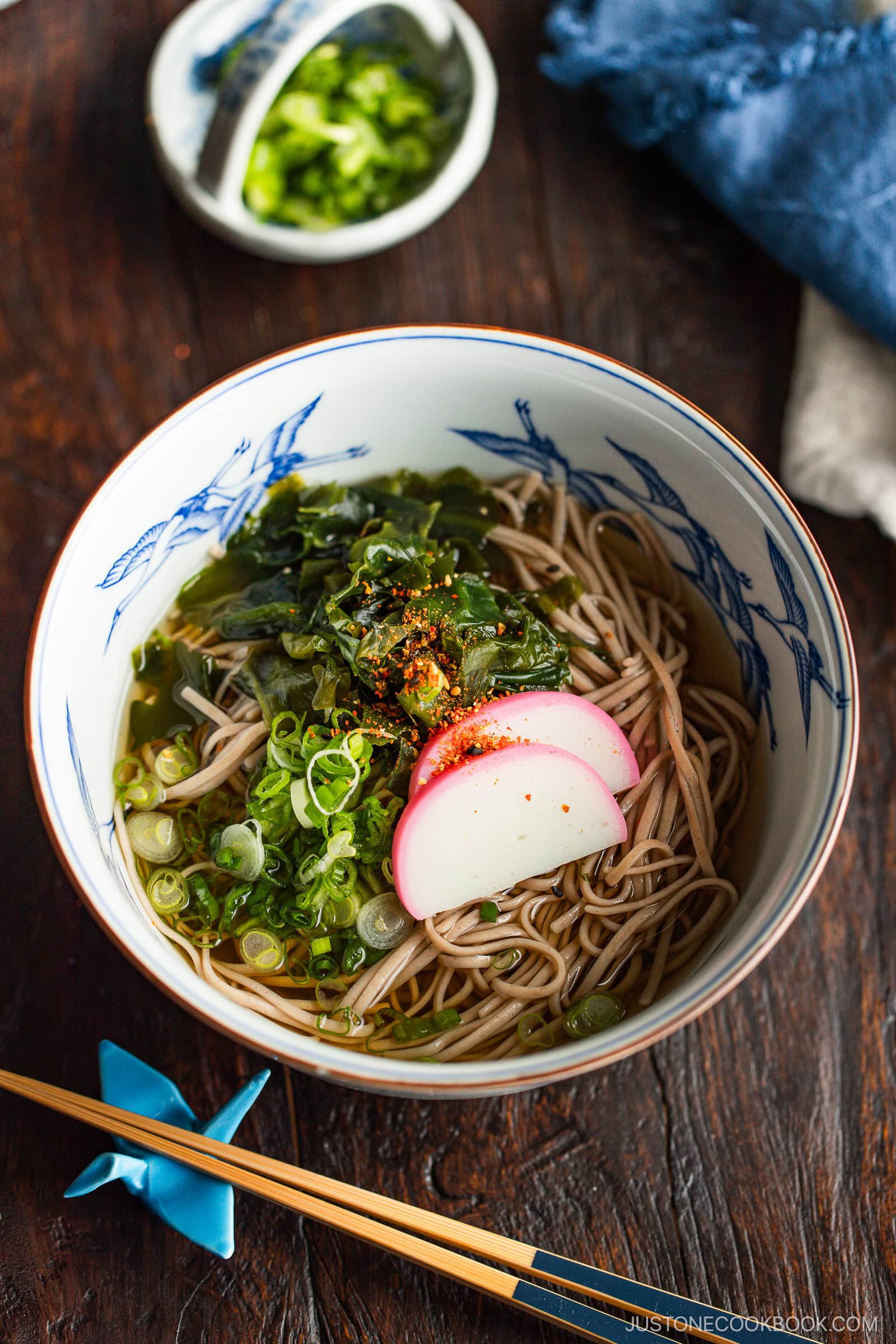 A bowl of soba noodle soup topped with sliced kamaboko, chopped green onions, seaweed, and a sprinkle of spices. Chopsticks and a dish of extra green onions are beside the bowl on a wooden table.