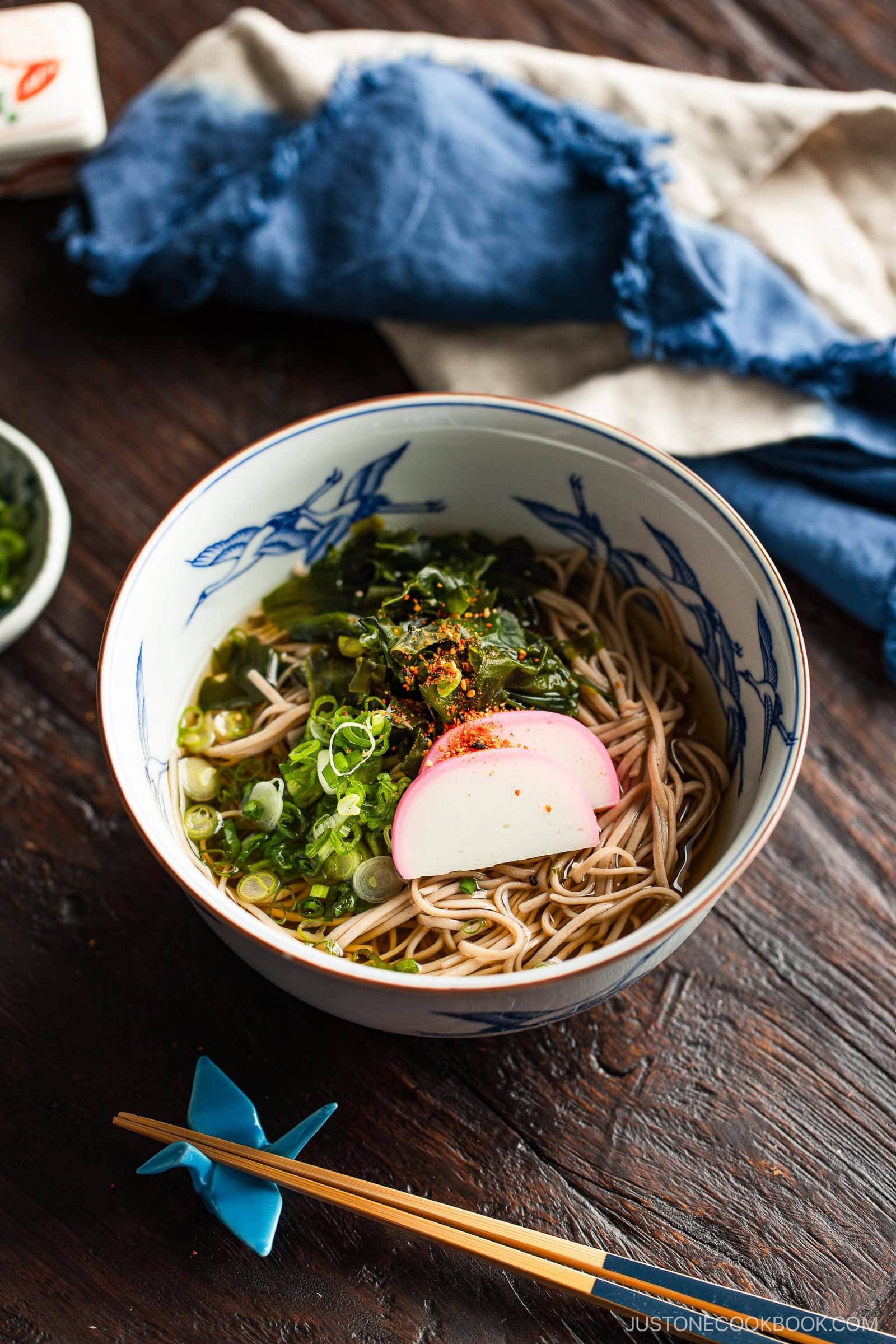 A bowl of soba noodle soup topped with sliced fish cake, green onions, seaweed, and seasoning, placed on a wooden table with chopsticks and a blue napkin nearby.