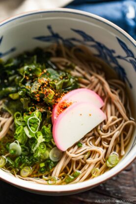 A bowl of soba noodle soup topped with green onions, seaweed, and three slices of pink and white fish cake, served in a blue and white patterned bowl.