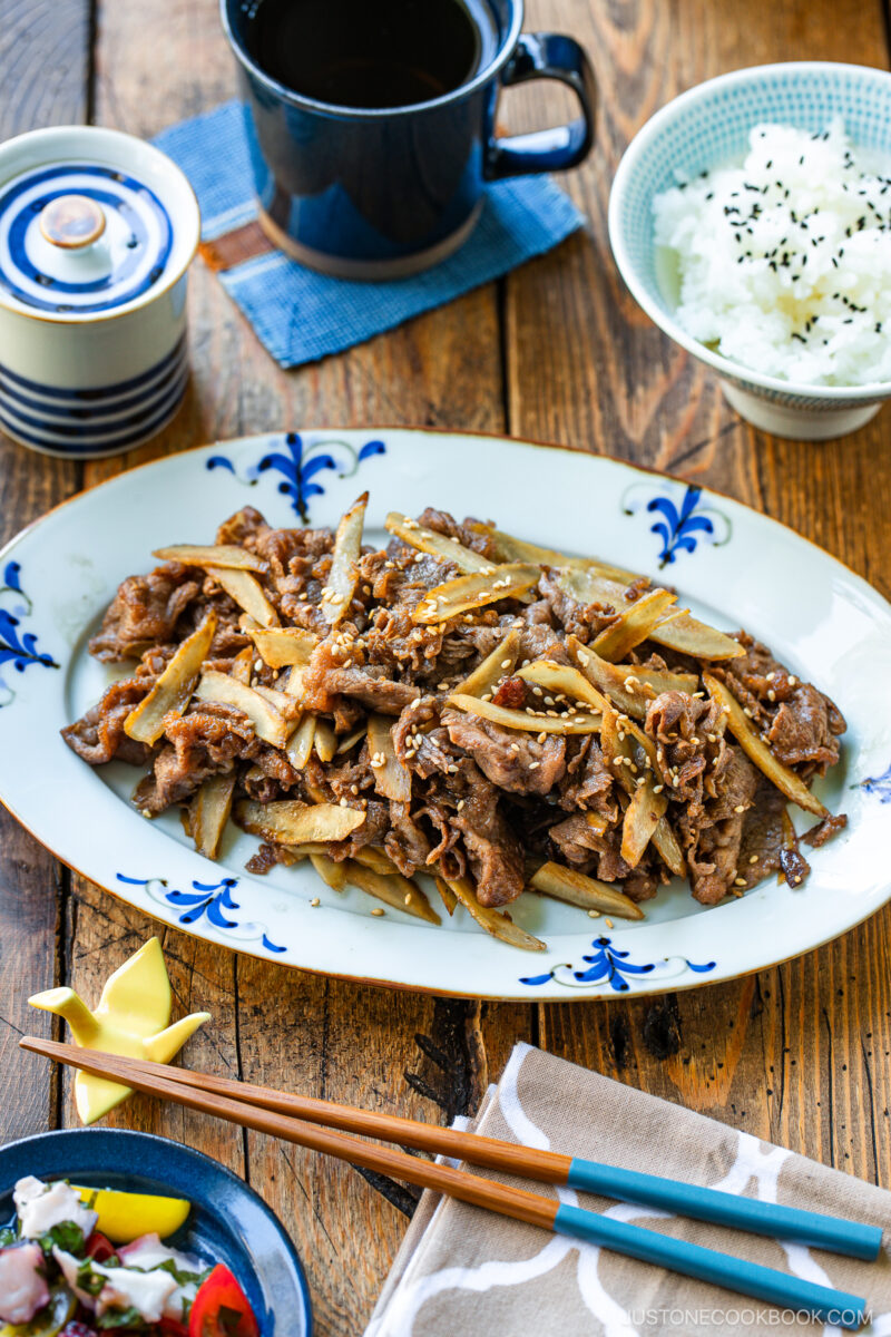 A plate of Beef and Gobo Stir Fry garnished with sesame seeds sits on a wooden table, accompanied by a bowl of white rice, a cup of tea, chopsticks, and a small salad.