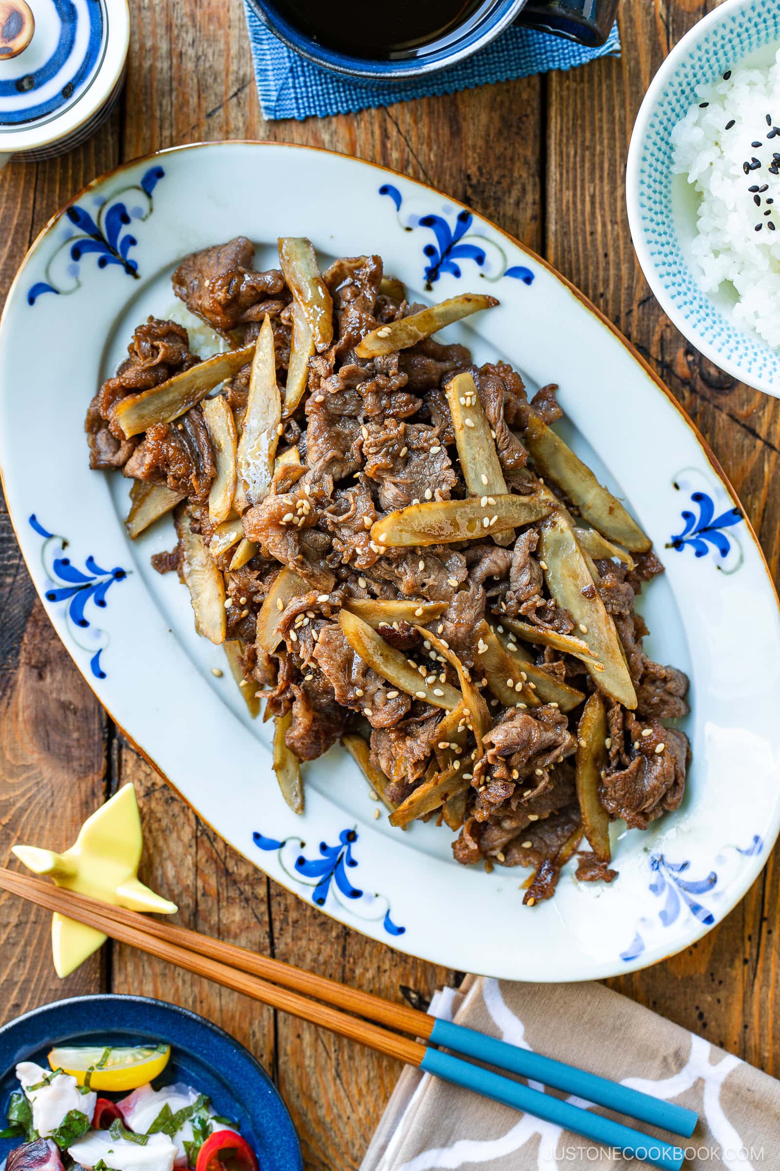 A blue-and-white oval plate holds Beef and Gobo Stir Fry&mdash;tender beef and burdock root garnished with sesame seeds. A bowl of rice, a small dish, and chopsticks with a blue holder are arranged nearby on a wooden table.